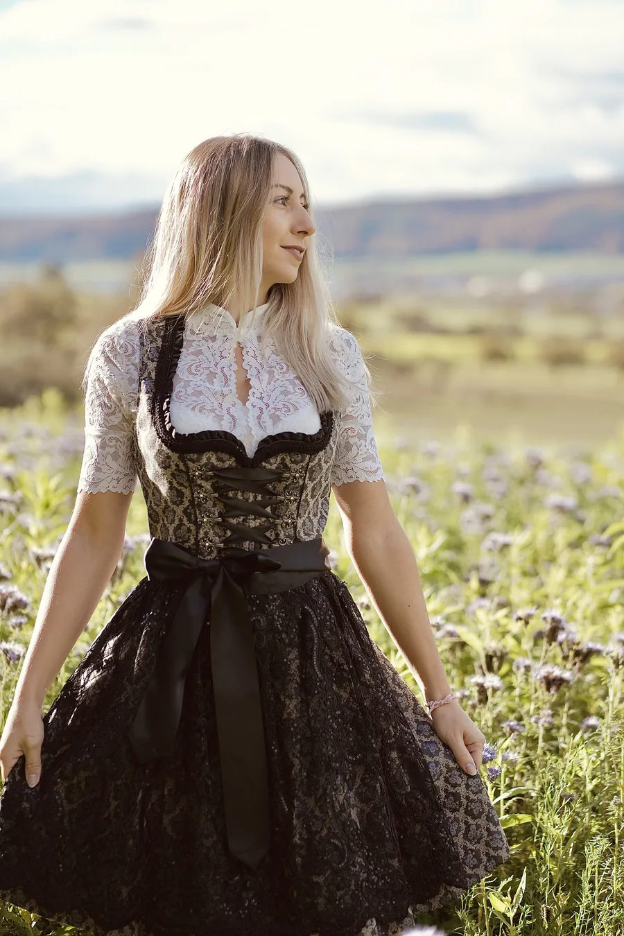 Frau im schwarzen Dirndl mit weißer Spitzenbluse hält den Rock auf einer Blumenwiese im Abendlicht